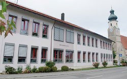 Das Bild zeigt die Straßenseite der Grundschule Haiming, rechts im Hintergrund ist der Kirchturm zu sehen.